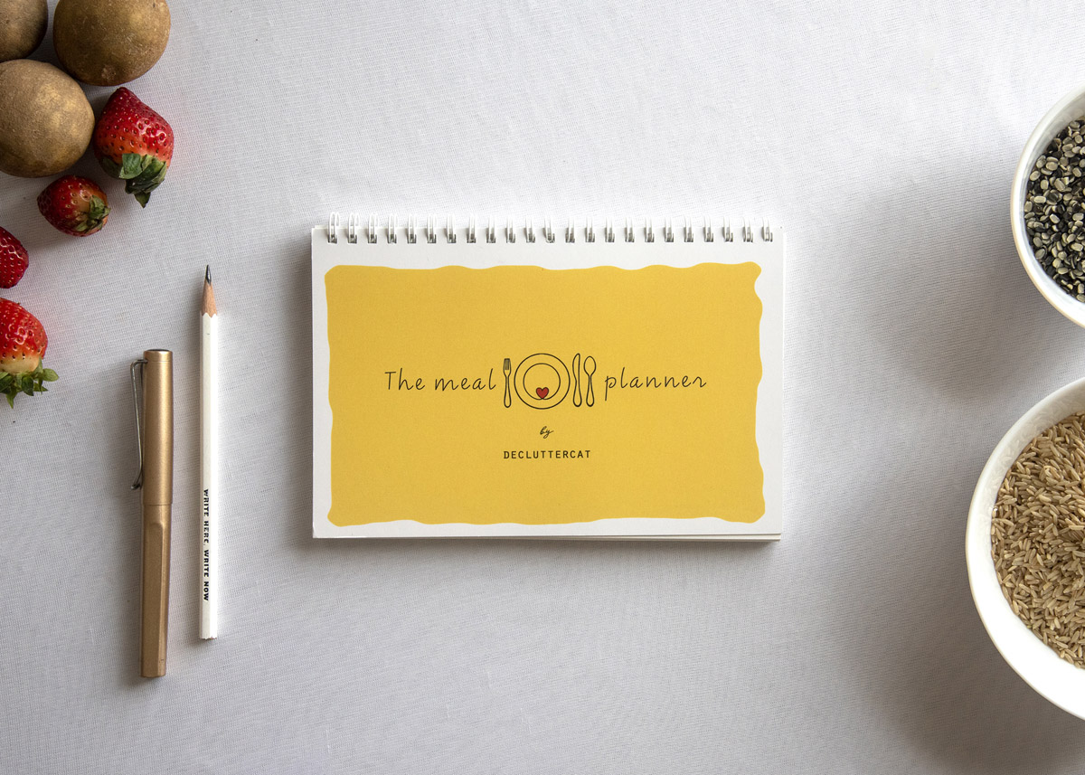 The Meal Planner by Decluttercat, a tool for mindful eating, shown on a white tablecloth with a pen, pencil, and healthy ingredients like strawberries and seeds, ready for festive meal planning